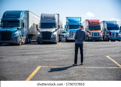 Slim Truck driver carries a box with purchases in his hands and going to his big rig semi ruck parked on the truck stop parking lot standing in row with another semi trucks with semi trailers