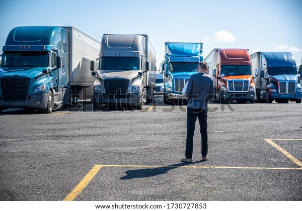 Slim\
Truck driver carries a box with purchases in his hands and going to\
his big rig semi ruck parked on the truck stop parking lot standing\
in row with another semi trucks with semi\
trailers