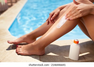 Slim woman applies suntan cream on legs sitting near swimming pool. Girl puts water resistant sunscreen lotion from a bottle on body on poolside deck. Skin burn and anti cancer care concept.
