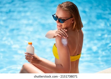 Slim woman applies suntan cream on shoulder sitting near swimming pool. Girl puts water resistant sunscreen lotion from a bottle on body on poolside deck. Skin burn and anti cancer care concept.