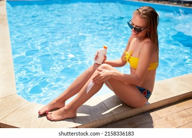 Slim woman applies suntan cream on legs sitting near swimming pool. Girl puts water resistant sunscreen lotion from a bottle on body on poolside deck. Skin burn and anti cancer care concept.