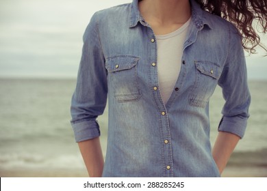 Slim Woman In A Denim Shirt And White T-shirt Stands On The Beach Against The Sea In Cloudy Weather. She Has Curly Hair. Her Hands In Her Pockets. Retro Colors. Closeup. Relaxed Mood.