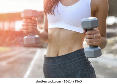 Slim woman with dumbells posing outdoors. She's doing exercises and wearing sport suit