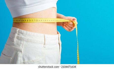 Slim woman measures her waistline with measuring tape. Healthy body shaping weight loss concept. Slim waist small belly in big white denim pants isolated over blue color background. Long web banner