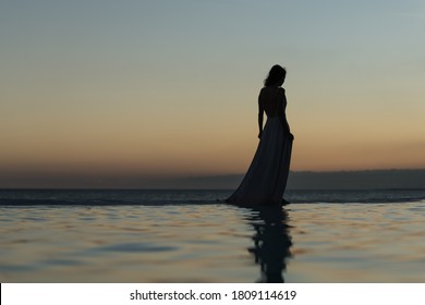 Slim woman silhouette walking on the pool edge against sunset sky