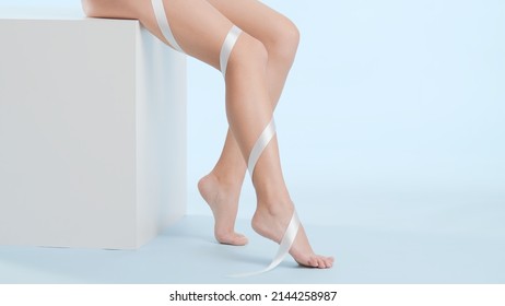 Slim woman with smooth legs sits on white cube podium on pale blue background. Silk ribbon wrapped leg | Leg skin care concept