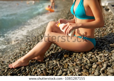 Slim woman tanned lower body in shape lying on pebble beach near sea waves and surf with sunblock cream bottle. Girl applies water resistant sunscreen lotion on legs relaxing in sun holiday.