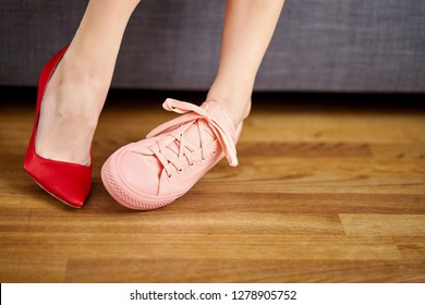 Slim Woman's Legs In Two Different Shoes With Red High Heels And Coral Sneakers On Gray Couch Background.