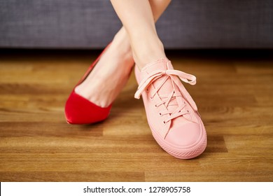 Slim Woman's Legs In Two Different Shoes With Red High Heels And Coral Sneakers On Gray Couch Background.