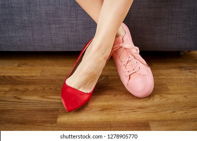 Slim Woman's Legs In Two Different Shoes With Red High Heels And Coral Sneakers On Gray Couch Background.