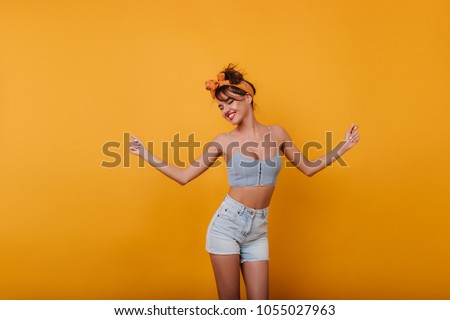 Slim young female model with vintage hairstyle posing on yellow background. Studio shot of winsome laughing girl dancing in denim shorts.