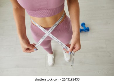 Slim young girl holding tape measure on her thin waist. Unrecognizable sporty woman showing her weightloss achievement after home workouts. Cropped close up, closeup. Beauty and weight loss concept
