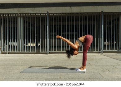 Slim young woman in modern tracksuit does uttanasana standing on mat against large building gate at training on sunny day