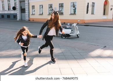 Slim Young Woman In Trendy Jeans And Leather Jacket Running Down The Street Holding Daughter's Hand. Joyful Long-haired Little Girl In Rock Style Outfit Having Fun With Mom Outside