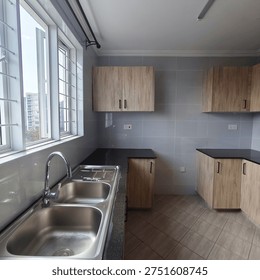 A slim-designed kitchen featuring wooden upper cabinets, a black countertop, and a modern faucet over a double stainless steel sink. The room has large windows with metal grilles