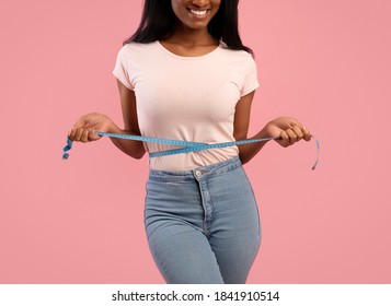 Slimming and dieting concept. Unrecognizable black woman measuring her waist during weight loss over pink studio background. Cropped view of young lady checking her body parameters