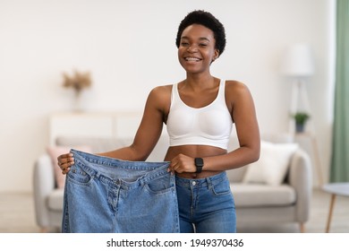 Slimming Motivation Concept. Skinny Black Female Showing Old Large Jeans After Successful Diet And Weight Loss Posing Smiling To Camera At Home. Weight-Loss Before And After Comparison