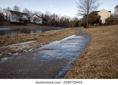 A slippery icy patch covers part of a walking path next to a peaceful pond in a quiet neighborhood of Ashburn, Virginia, on a cold winter day.