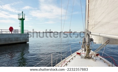 Sloop rigged yacht leaving port at sunset. Cargo terminal and ships in the background. Baltic sea. Nautical vessel, transportation, service, sport, recreation concepts