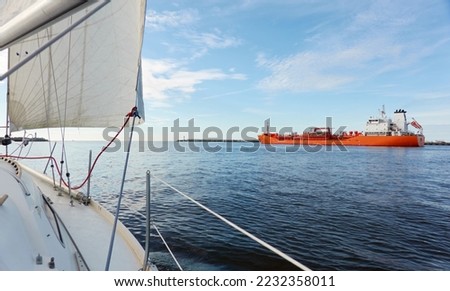 Sloop rigged yacht leaving port at sunset. Cargo terminal and ship in the background. Baltic sea. Nautical vessel, transportation, service, sport, recreation concepts