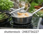 Slotted spoon takes golden deep-fried chicken pieces from a steaming pan with bubbling boiling oil, outdoor kitchen stove surrounded by potted herbs and vegetables, copy space, selected focus 