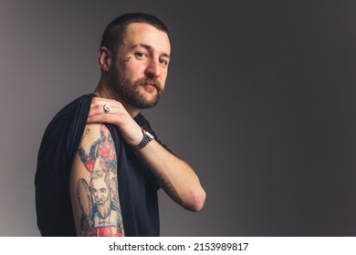 Slouchy gangster man in black t  shirt showing to the camera tattoos his shoulder over dark gray background  High quality photo