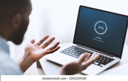 Slow computer software concept. Indignant african american guy looking at laptop with loading screen. Over shoulder shot, selective focus on device
