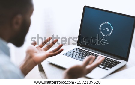 Slow computer software concept. Indignant african american guy looking at laptop with loading screen. Over shoulder shot, selective focus on device