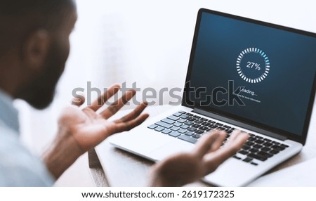 Slow computer software concept. Indignant african american guy looking at laptop with loading screen. Over shoulder shot, selective focus on device