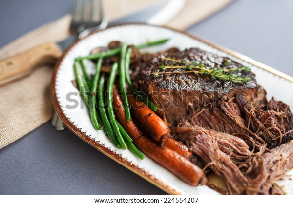 Slow cooked pot roast with\
carrots, green beans, onions, garlic and gravy on a white porcelain\
platter with gold rim and serving fork against a gray tablecloth.\
