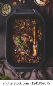 Slow Cooked Venison Roast In Black Cast Iron Pan, Top View. Braised Leg Of Deer With Bone And Roasted Vegetables In Pot On Dark Rustic Kitchen Table Background, Top View