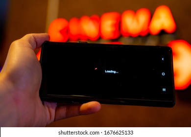 Slow Internet, Video Load And Download Speed. Watching Movie Online. Loading Icon On Screen.Hand With Black Empty Screen Mobile Phone . Background Red Wall With Cinema Signate.