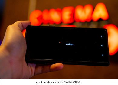 Slow Internet, Video Load And Download Speed. Watching Movie Online. Loading Icon On Screen.Hand With Black Empty Screen Mobile Phone . Background Red Wall With Cinema Signage.