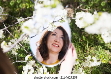 Slow Life. Enjoying The Little Things. Dreaming Of Spring. Middle Aged Woman Holds And Looks In Her Reflection In A Round Mirror, Relax In The Wind In Flowering Spring Garden. Burnout Syndrome