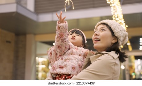 Slow Motion Of Cute Curious Asian Toddler Baby Girl Raising Up Hand Trying To Touch Outdoor Christmas Lights In Her Mother’s Arms Outside An Urban Shopping Mall