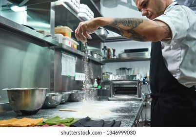 Slow Motion. Handsome Young Chef With Black Tattoos On His Arms Pouring Flour On Kitchen Table Before Making Pasta