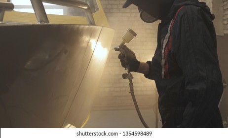 In Slow Motion A Professional Man (guy) In A Garage (service) In Working Clothes Dressed In A Sweater Sets Up A Paint Gun (spray Gun) And Starts Painting (putting A Layer Of Ceramics) A Yacht.