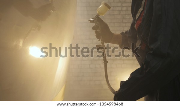 In slow motion a professional man (guy) in a\
garage (service) in working clothes dressed in a sweater sets up a\
paint gun (spray gun) and starts painting (putting a layer of\
ceramics) a yacht.