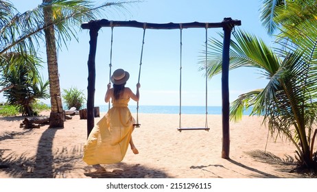 Slow motion of unrecognizable travel woman on wooden swing on tropical sandy beach with blue sea and palm trees. Female traveler on Phuket island, Thailand. Vacation and summer holidays concept. 