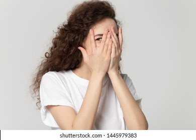 Sly 20s Girl Wearing White T-shirt Hiding Her Face Behind Hands Peeps Through Fingers Posing Isolated On Gray Studio Background. Crafty Look Of Curious Woman Feels Afraid Or Shy Peeking Concept Image