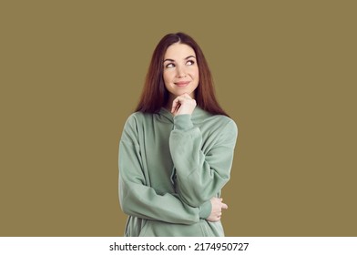 Sly Young Woman With Smile Ponders Over Idea, Looking Aside On Khaki Background. Joyful Caucasian Woman In Casual Hoodie Touching Her Chin And Looking At Upper Right Corner With Sly Expression.