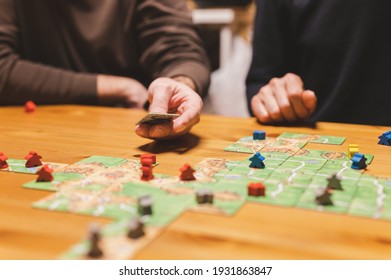 Smakhtino, Russia, December 2020: Two Men Friends Have Fun Playing The Carcassonne Board Game Late In The Evening Or At Night. Male Hands And Game Cards And Chips On Table