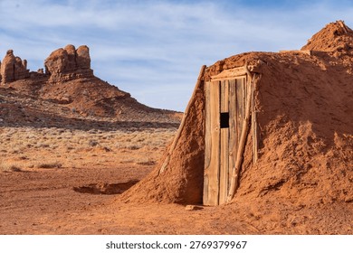 A small adobe structure sits on the ground in a desert area. Surrounding the structure are dry grasses and notable rock formations. The sky shows scattered clouds above.