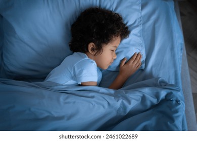 A small African American child is laying comfortably in a bed covered with a blue comforter. Boy appears to be resting peacefully, with eyes closed and hands relaxed by his side - Powered by Shutterstock