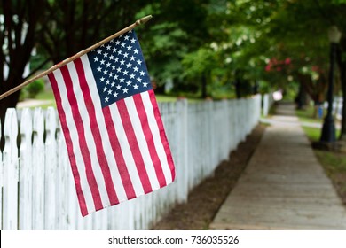 Small American flag hangs from a picket fence along the sidewalk in a rural town.