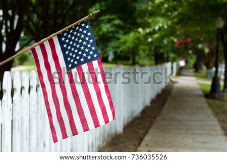Small American flag hangs from a picket fence along the sidewalk in a rural town.