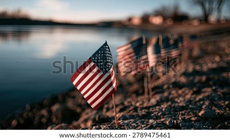 Small American flags planted in a row along a rocky shore next to a body of water during a sunset.
