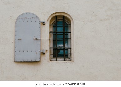 A small arched window is set into a smooth beige stucco wall, framed by a slightly recessed border that creates subtle depth. The window features multiple vertical and horizontal black metal bars forming a grid pattern across the glass. Behind the bars, a sheer curtain in muted blue tones is partially visible, softening the interior view. To the left of the window, a hinged metal shutter is mounted on the wall, painted in a light gray color and reinforced with evenly spaced rivets along its edges. The shutter is open, revealing its curved top that mirrors the shape of the window arch. A thin metal rod extends from the shutter toward the wall, suggesting a mechanism for securing it in place. The composition is minimal, with the plain textured surface of the wall occupying most of the frame, emphasizing the contrast between the neutral background and the darker structural elements. Soft natural lighting casts gentle shadows, highlighting the textures of the plaster and the matte finish of the metal fixtures.