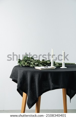 Small artificial tree, notepad, glasses and unlit candles on a round table with a black rough tablecloth. Cropped, side view.