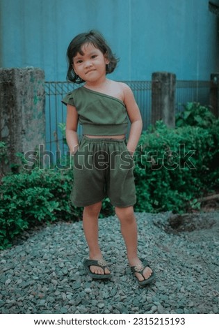 a small Asian girl wearing a one-shoulder sleeveless slash neck summer blouse is standing on a pile of rocks while posing with both hands tucked into the pockets of her pants